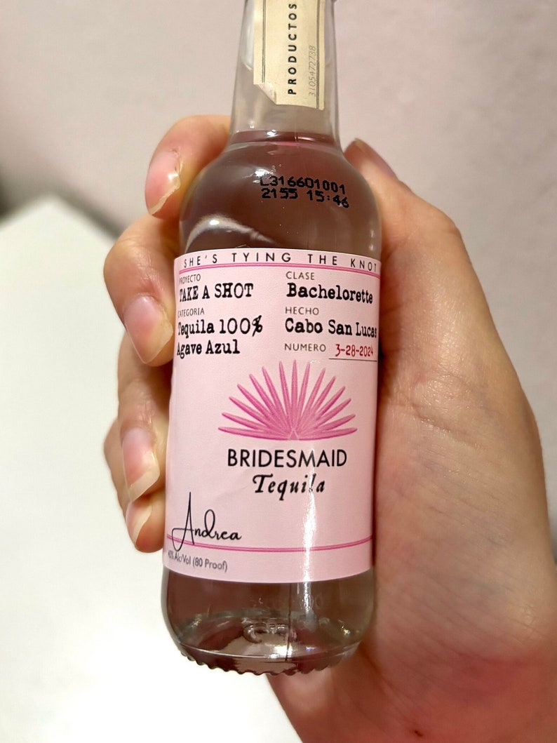May include: A pink and white bottle of Bridesmaid Tequila with the text "Take a Shot Tequila 100% Agave Azul" and "She's Tying the Knot Bachelorette Cabo San Lucas 3-28-2024" on the label.