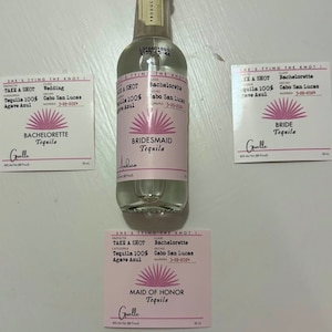 May include: Four small pink and white labels with the text "She's tying the knot!" and "Take a shot Tequila 100% Agave Azul" with a pink sunburst design. The labels are for a wedding, bachelorette party, bridesmaid, and maid of honor. The labels are attached to a clear glass bottle of tequila.