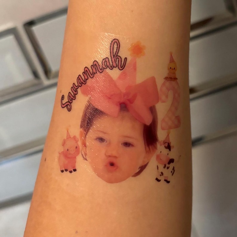 May include: A temporary tattoo with a child's face, a pink bow, and the name "Savannah". The design includes the number "2", a pig, and a cow, all in a colourful style.