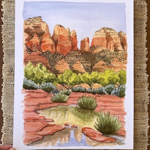 May include: Watercolor painting of a desert landscape featuring red rock formations, green shrubbery, and a small pool of water. The sky is a light blue, and the artwork is signed in the lower left corner. The painting is on white paper.