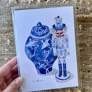 May include: A watercolor print featuring a blue and white ginger jar and a nutcracker. The jar has a bird and floral design. The nutcracker is in a matching blue and white uniform. The print is signed "g. Leman".