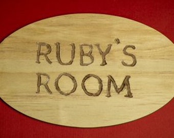 Puede incluir: Un letrero ovalado de madera con el texto "RUBY'S ROOM" grabado en la madera.