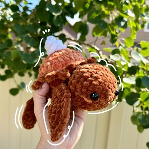May include: A handmade, plush turtle in shades of brown and orange. The turtle has a textured, velvety appearance, with a white pom-pom on its back. The turtle's eyes are black, and it has a smiling expression. The turtle is a soft toy.