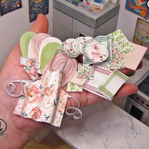 May include: A set of miniature dollhouse kitchen accessories, including a floral print apron, plates, and napkins. The items are made of paper and are designed in a pink and green floral pattern. The set is labeled "1:12 scl."
