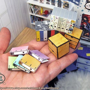 May include: Miniature dollhouse office supplies, including colorful binders, wooden drawers, and file folders. The binders are in various colors, such as pink, yellow, and green. The scale is 1:12.