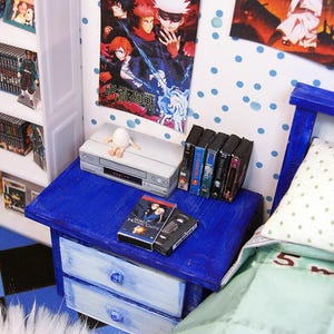 May include: Miniature bedroom scene with a blue nightstand, a VCR, and several VHS tapes. A poster with Japanese text hangs on the wall. The scene includes a bed with a green and white patterned comforter. The scale is 1:12.