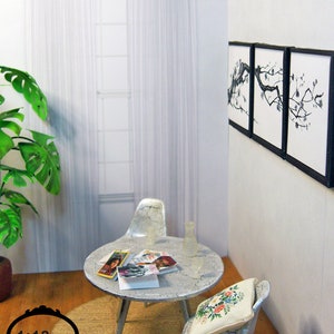 May include: Miniature dollhouse living room scene with a round table, two chairs, and a window with sheer curtains. The table has magazines and a vase. Artwork hangs on the wall. The scene is set on a wooden floor with a rug.