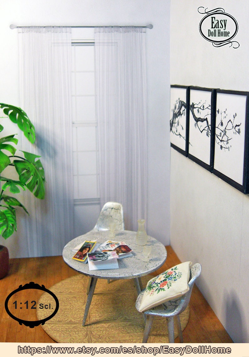 May include: Miniature dollhouse living room scene with a round table, two chairs, and a window with sheer curtains. The table has magazines and a vase. Artwork hangs on the wall. The scene is set on a wooden floor with a rug.