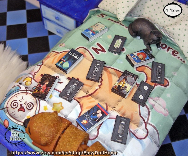 May include: Miniature VHS tapes and movie boxes are scattered on a patterned blanket with a teddy bear. The boxes feature movie titles like "Totoro" and "Howl's Moving Castle." A small black cat figurine sits on the blanket. The scale is 1:12.