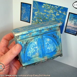 May include: A miniature blue cabinet with ornate detailing and a floral pattern on the doors. The cabinet is part of a dollhouse set, with matching artwork on the wall. The scale is 1:12.