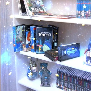 May include: Miniature white bookshelf with various items. Includes magazines, VHS tapes, and books, including "My Neighbor Totoro." The shelf also features small figurines and a collection of miniature books. The scale is 1:12.