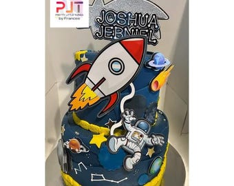 May include: A two-tiered blue cake decorated with a rocket, astronaut, stars, and planets. The cake is topped with a silver and black sign that says "Joshua Jebniel".