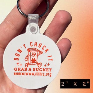 May include: A round white keychain with a metal ring. The front features an orange graphic of a cartoon bucket with a biohazard symbol, the text DON'T CHUCK IT, GRAB A BUCKET, and the website NHHRC.ORG. The keychain measures 2 inches by 2 inches.