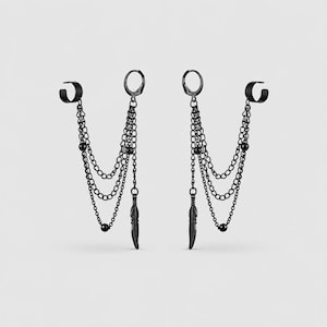 May include: A pair of matte black earrings featuring a hoop and a cuff connected by three draped chains. Each earring includes small round beads and a single feather-shaped charm hanging from the chains, creating a layered, dangling design.