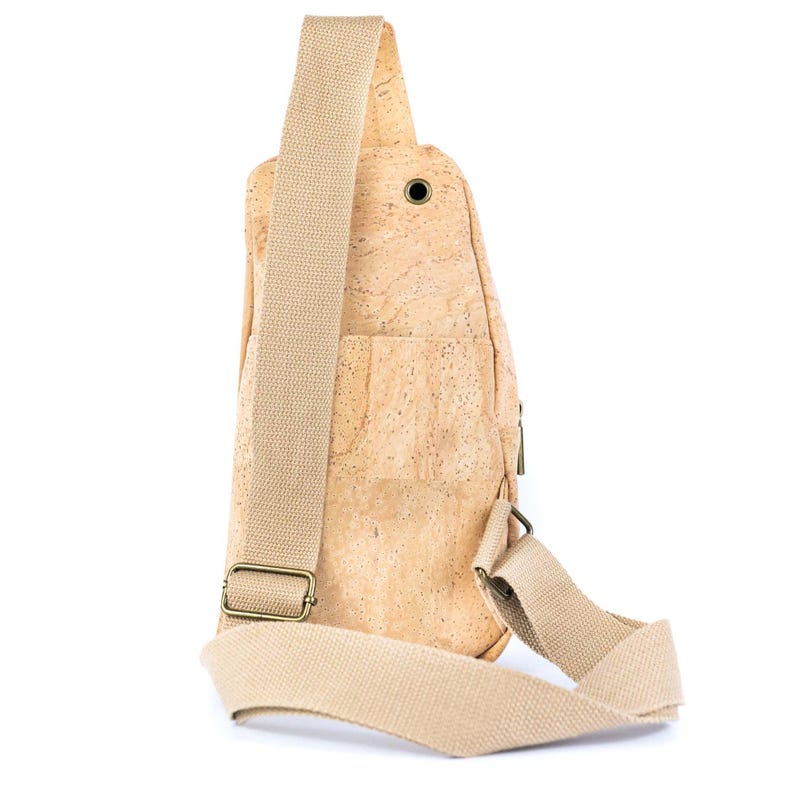 May include: A tan cork sling bag featuring a textured natural surface, a single wide beige adjustable fabric shoulder strap, a metal buckle, and a small circular metal grommet detail on the upper front section of the main body.