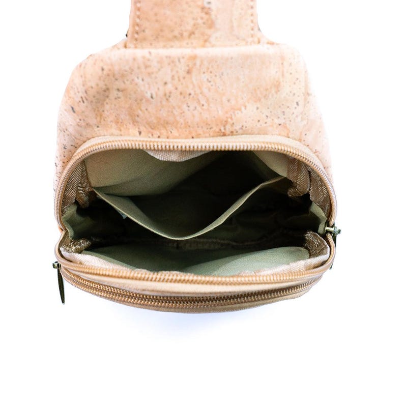 May include: A light tan cork sling bag with a speckled texture, featuring a zippered main compartment that is open to reveal a beige fabric interior lining with a single slip pocket and a secondary zippered front pocket with matching metal pulls.
