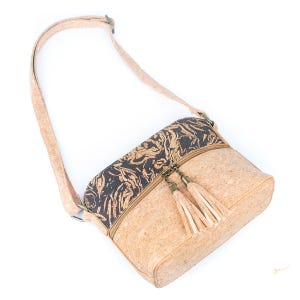 May include: A light brown crossbody bag made of cork material. The bag features a black and brown patterned top section, a zipper closure, and two beige tassels. The adjustable strap is also light brown.