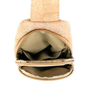 May include: A light brown cork sling bag with a natural speckled texture. The bag features two zippered compartments with tan fabric lining, a vertical strap attachment at the top, and a structured shape designed for carrying small personal items.