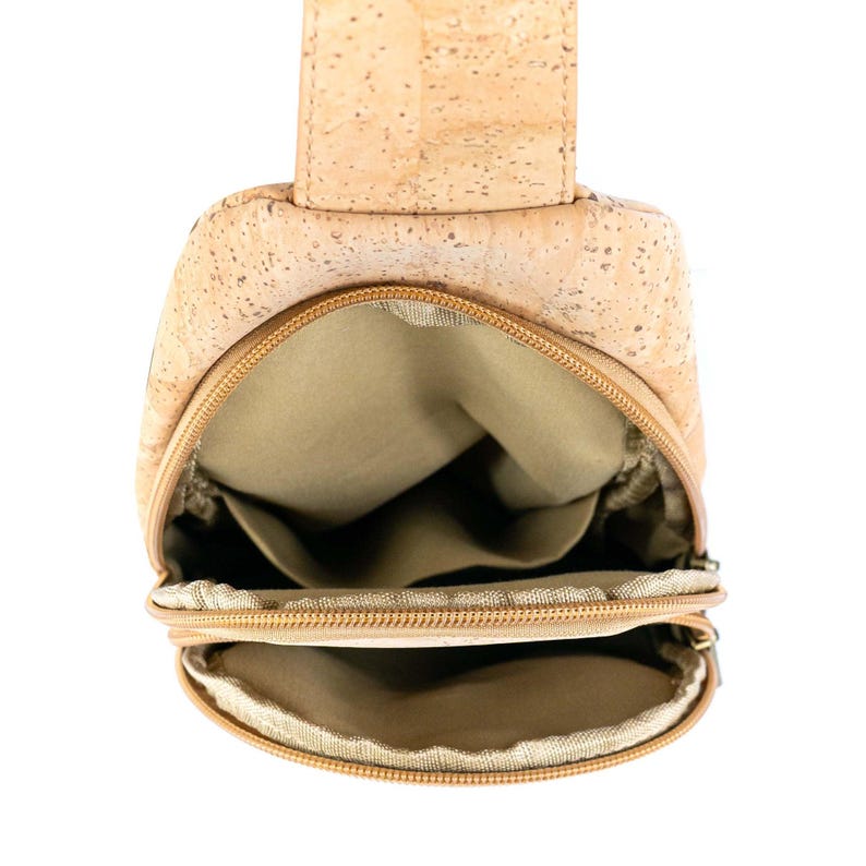 May include: A light brown cork sling bag with a natural speckled texture. The bag features two zippered compartments with tan fabric lining, a vertical strap attachment at the top, and a structured shape designed for carrying small personal items.