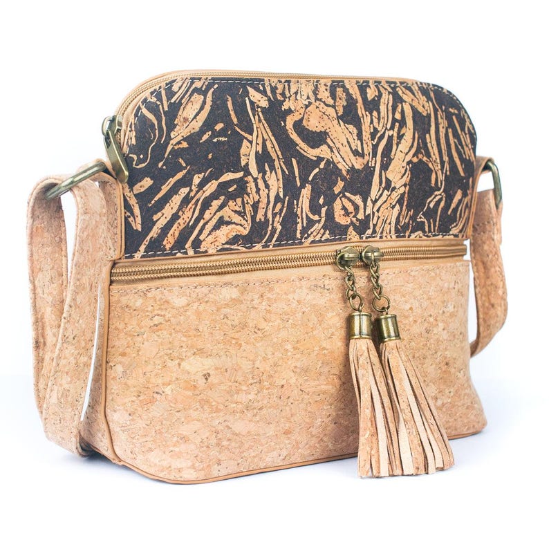 May include: A crossbody bag made of cork material. The bag features a light brown base with a dark brown and tan patterned top section. It has a zipper closure with decorative tassels and a long adjustable strap. The bag is designed with a rectangular shape.