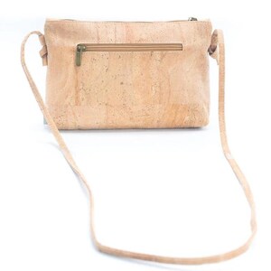 May include: A light brown crossbody bag made of cork material. The bag has a rectangular shape with a zipper closure and a long, thin strap. The bag's texture is visible, and the hardware is a brass color.