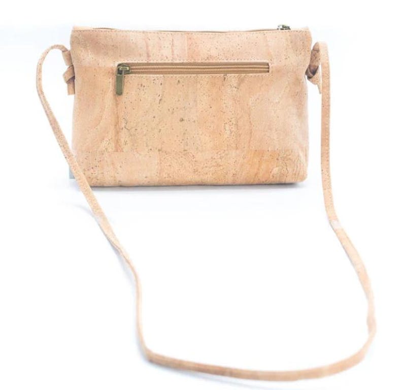 May include: A light brown crossbody bag made of cork material. The bag has a rectangular shape with a zipper closure and a long, thin strap. The bag's texture is visible, and the hardware is a brass color.