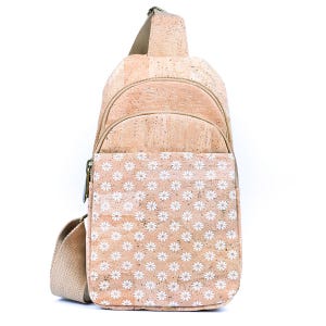 May include: A tan cork sling backpack featuring a natural speckled texture and a repeating white daisy floral pattern. The bag includes a top zippered compartment, a front zippered pocket, and a wide, adjustable beige fabric shoulder strap.