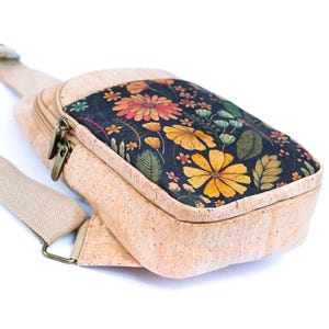 May include: A crossbody bag crafted from light tan cork material featuring a rectangular front panel with a black background and a vibrant floral print in shades of yellow, orange, and green. The bag includes a beige fabric strap and metal zippers.