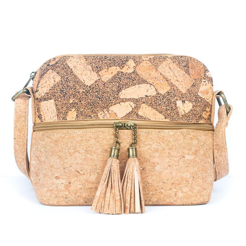 May include: A rectangular crossbody bag made of cork material. The bag features a light brown color with a textured pattern on the top section. It has a zipper closure and two decorative tassels hanging from the zipper. The bag has a long adjustable strap.