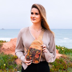May include: A tan cork crossbody sling bag featuring a front pocket with a dark floral pattern of orange, yellow, and green leaves and flowers. The bag has an adjustable tan strap and a zipper closure, designed to be worn across the chest or back.