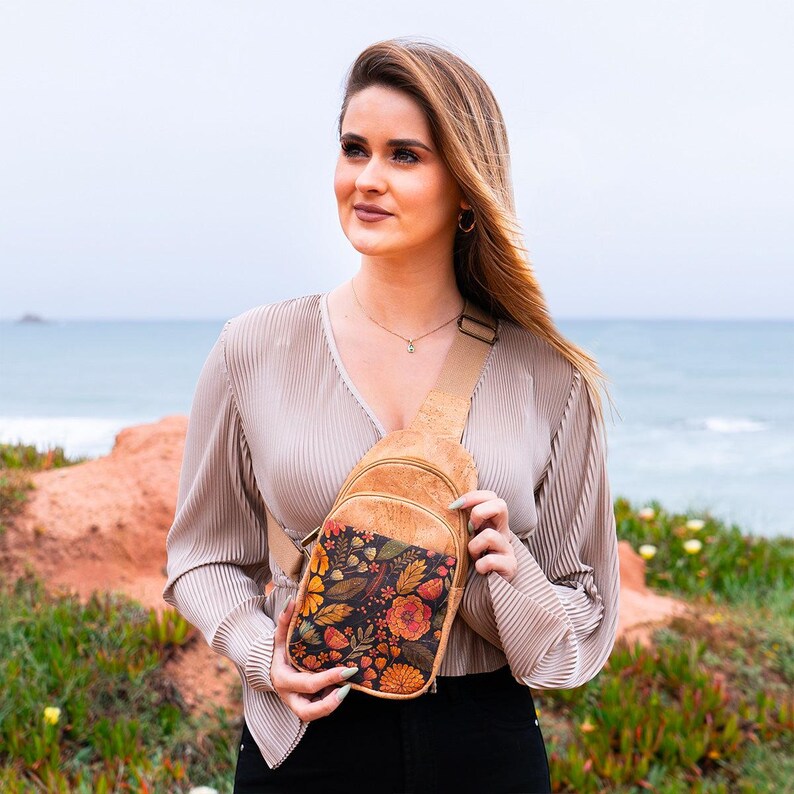 May include: A tan cork crossbody sling bag featuring a front pocket with a dark floral pattern of orange, yellow, and green leaves and flowers. The bag has an adjustable tan strap and a zipper closure, designed to be worn across the chest or back.