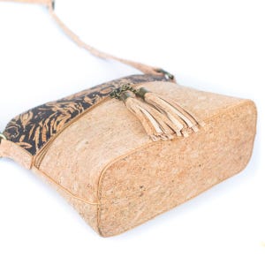 May include: A light brown cork crossbody bag with a black patterned accent on the top. The bag features a zipper closure and two beige tassels. The strap is thin and adjustable. The bag is made of cork material.