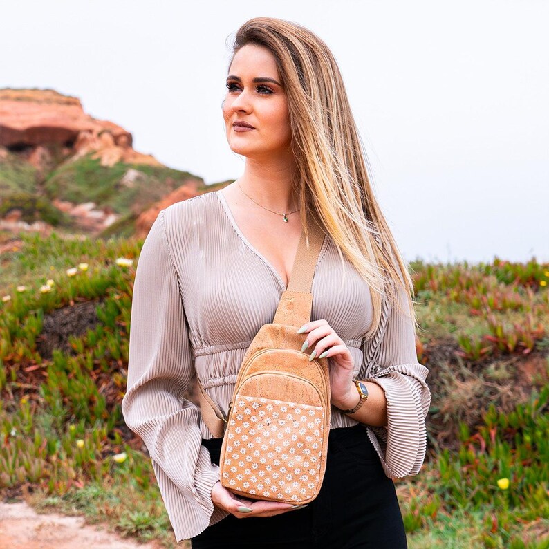 May include: A tan cork sling backpack featuring a front pocket with a white daisy floral pattern, a single adjustable shoulder strap, and a zipper closure, constructed from natural cork material with a textured finish and gold-tone hardware.