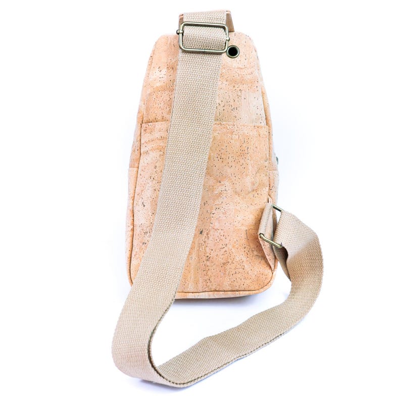 May include: A sling bag crafted from light tan cork material with a speckled texture. The bag features a wide, adjustable beige woven fabric strap with bronze-toned metal hardware, including a slider and a small circular grommet at the top.