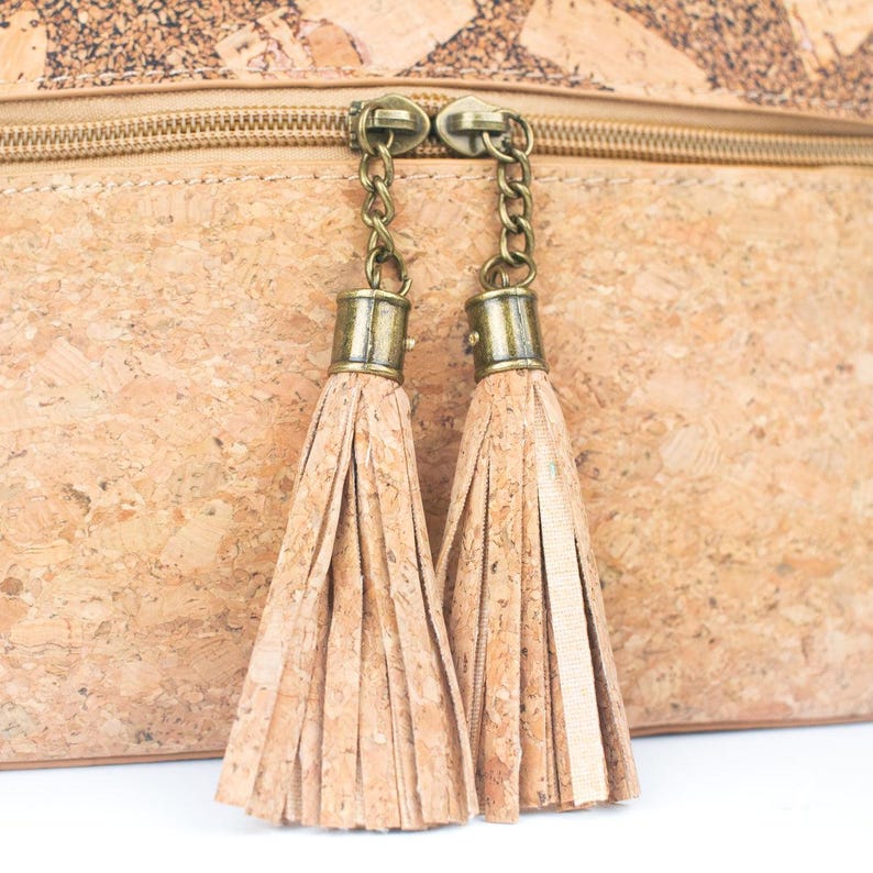 May include: Close-up of a cork bag with a zipper and two decorative tassels. The bag is light brown with a textured surface. The tassels are made of the same material and are attached to the zipper with small chains.
