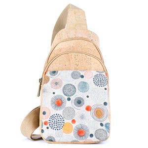 May include: A sling backpack crafted from natural tan cork material, featuring a front pocket adorned with a pattern of various circular designs in shades of orange, blue, gray, and white. The bag includes a beige woven strap and metal zippers.