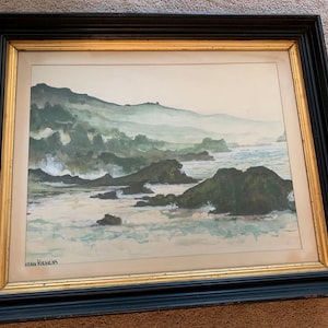 May include: Watercolor painting of a coastal landscape with a black and gold frame. The artwork depicts a misty shoreline with waves crashing against dark rocks. The artist's signature is visible in the lower left corner.