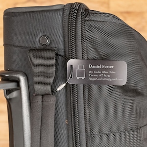 May include: Black luggage tag with a silver luggage icon and the text "Daniel Foster", "5615 Cedar Glen Drive", "Tucson, AZ 85747", and "FingerCraftsCo@gmail.com".