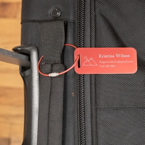 May include: Red luggage tag with a mountain design and the text "Kristina Wilson", "FingerCraftsCo@gmail.com", and "(123) 456-7890".