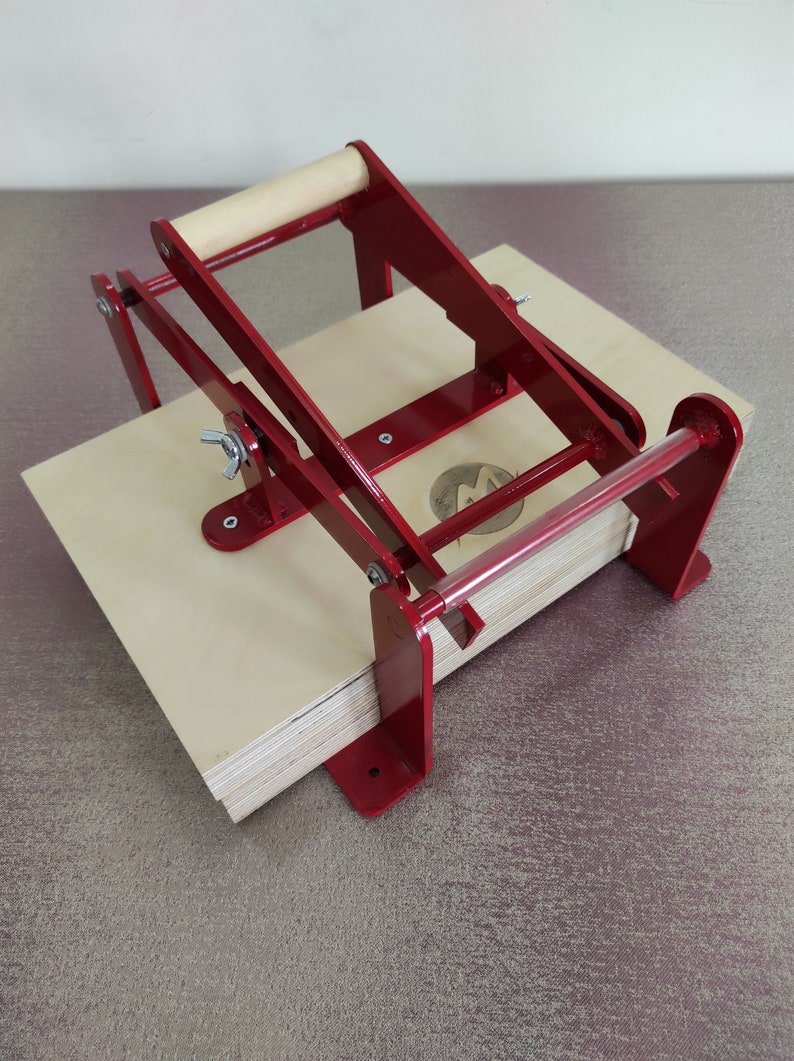 May include: A red metal book press with a wooden handle. The press is open and has a stack of plywood boards inside.