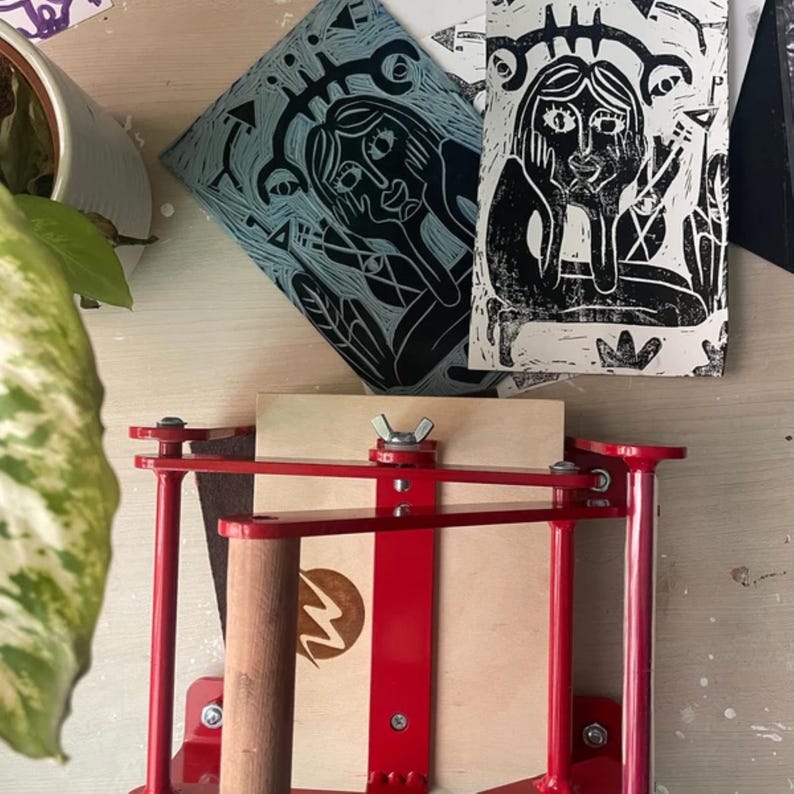 May include: A red printing press with a wooden handle and a wooden platform. Two black and white prints of stylized figures are on the platform.