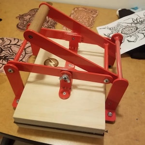 May include: A red metal printing press with a wooden handle. The press is made of metal and has a wooden base. The press is used to create prints from woodblocks or other materials.