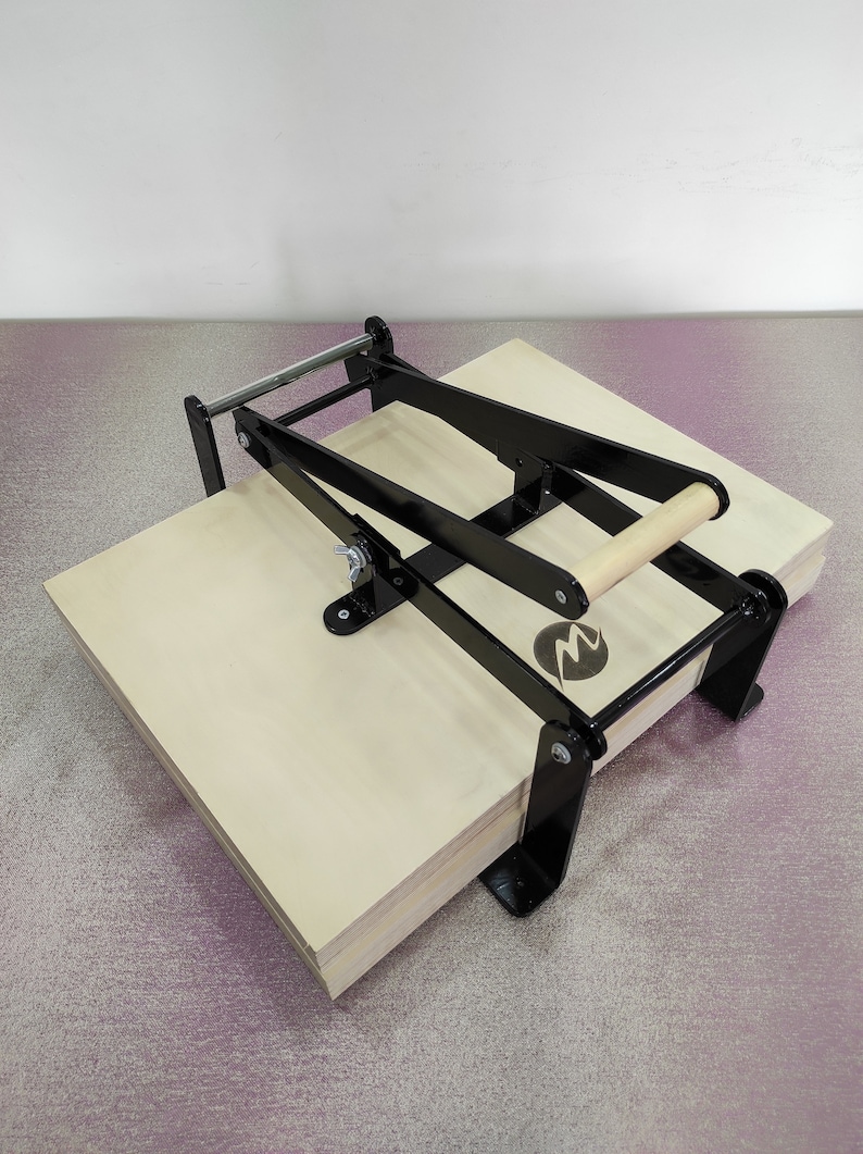 May include: A black metal book press with a wooden handle. The press is open and has a stack of plywood boards inside.