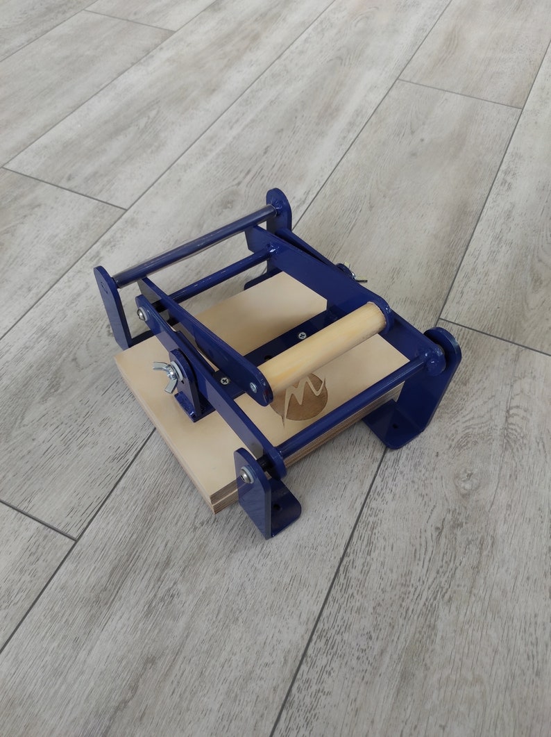 May include: A blue metal press with a wooden handle and a wooden base. The press has a cutout in the shape of a letter "M".
