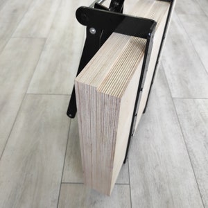 K&ouml;nnte beinhalten: Eine schwarze Metallklemme h&auml;lt zwei Sperrholzplatten zusammen. Das Sperrholz ist hellbraun und hat eine Holzmaserung.