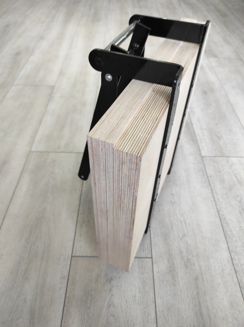 K&ouml;nnte beinhalten: Eine schwarze Metallklemme h&auml;lt zwei Sperrholzplatten zusammen. Das Sperrholz ist hellbraun und hat eine Holzmaserung.
