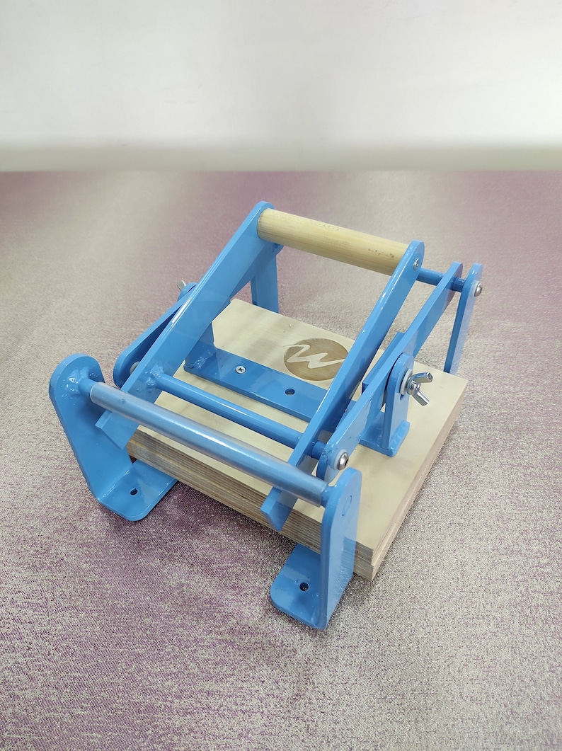 May include: A blue metal book press with a wooden handle. The press has a metal bar that runs across the top and a wooden platform that sits on top of the bar. The press is designed to hold books together while they are being glued or bound.