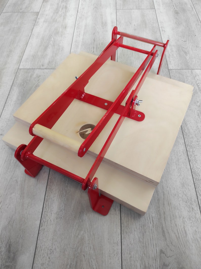 May include: A red metal press with a wooden handle, used for pressing items like fabric or paper. The press is sitting on top of two wooden boards.