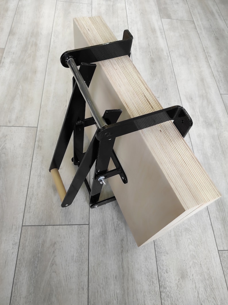 K&ouml;nnte beinhalten: Eine schwarze Metallklemme mit einem Holzgriff, die zum Halten eines Stapels Sperrholzplatten verwendet wird. Die Klemme ist zusammengeklappt und das Sperrholz lehnt an einer Wand.