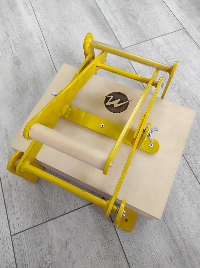 May include: A yellow metal press with a wooden handle, mounted on a wooden base. The press has a metal lever and a metal latch. The base has a circular logo with the letter "W" inside.